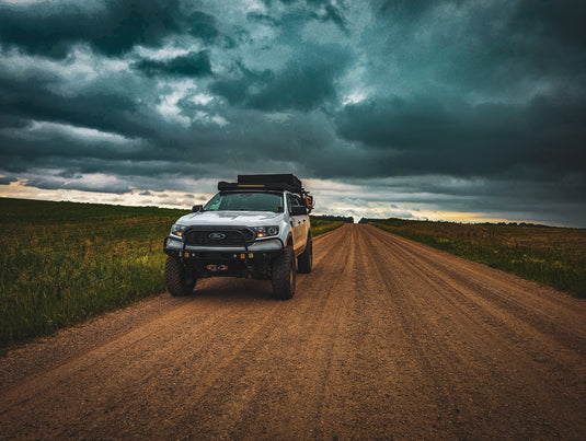 White truck with roof rack on a dirt road under a dark, stormy sky.